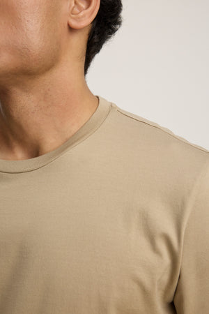 A close-up shows a person wearing the ALFIE TEE by Velvet by Graham & Spencer—a beige tailored fit crewneck—featuring only the lower face, neck, and upper torso against a plain background.