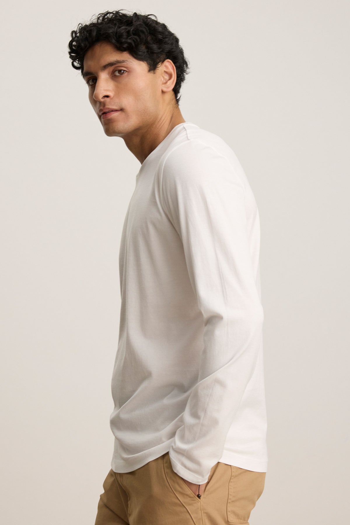 A man wearing the Velvet by Graham & Spencer ALFIE TEE in a tailored fit and tan pants stands sideways, facing the camera against a plain background.-39450513080513