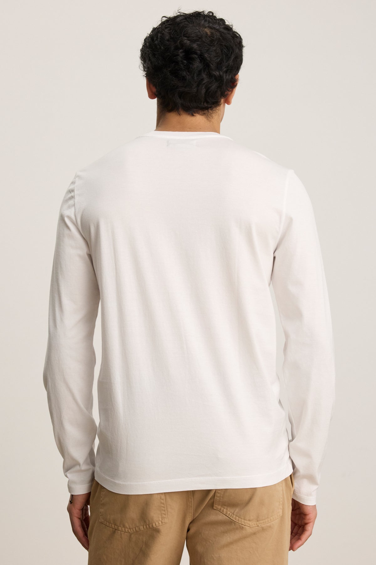 A person with short curly hair faces away, wearing the ALFIE TEE by Velvet by Graham & Spencer in white, paired with beige pants, against a plain background.-39450513113281