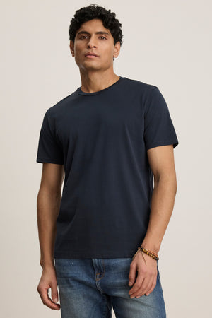 A man stands facing forward in the FRANKIE TEE by Velvet by Graham & Spencer—a plain navy blue crew neck t-shirt—paired with blue jeans against a neutral background.