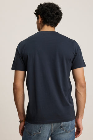 A man faces away from the camera, wearing the FRANKIE TEE by Velvet by Graham & Spencer—a plain dark blue crew neck t-shirt—paired with blue jeans against a light background.