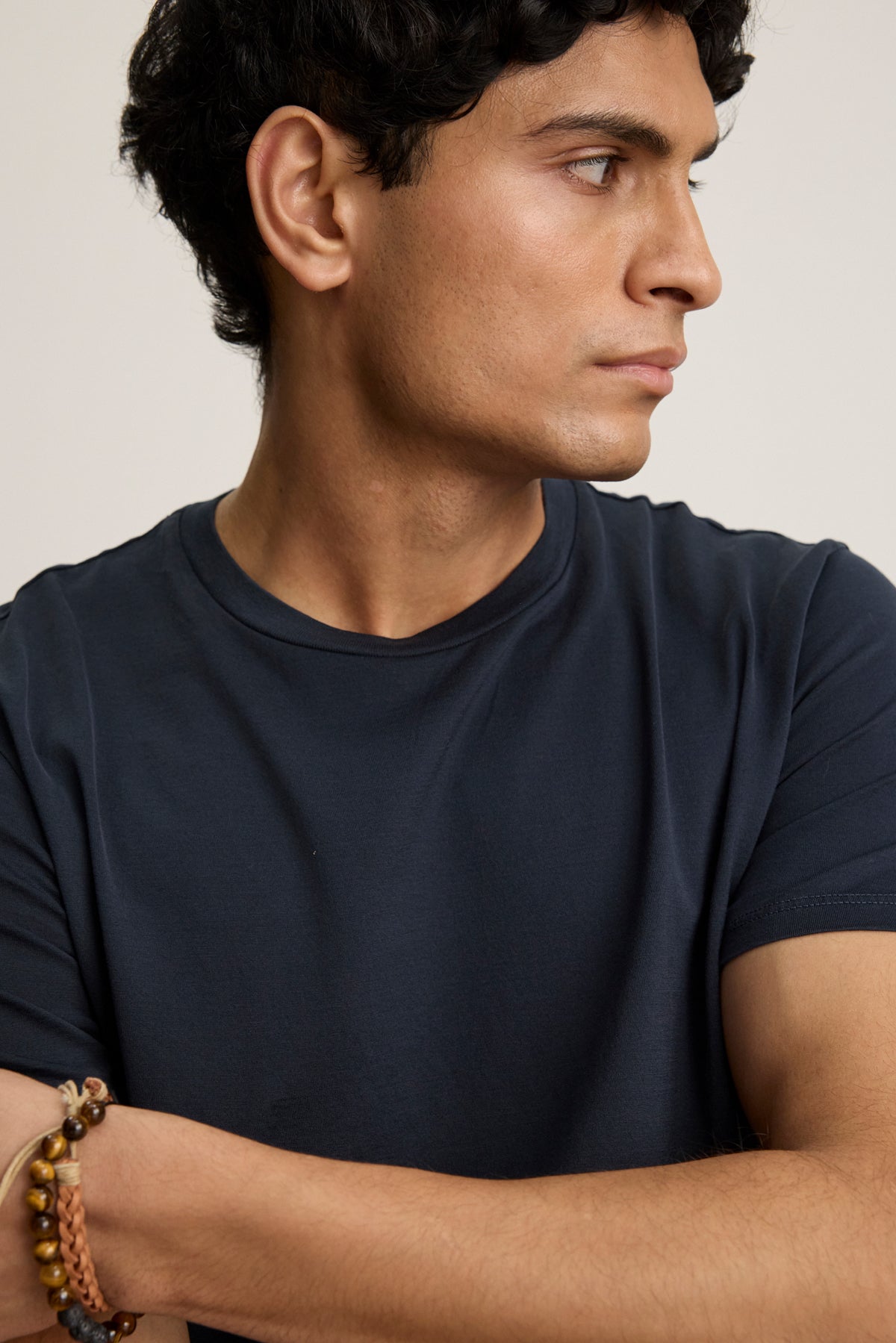 A man with short curly hair wears the FRANKIE TEE by Velvet by Graham & Spencer—a dark navy mercerized cotton crewneck—paired with beaded bracelets, looking right with his arms crossed.-39450522190017