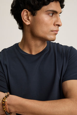 A man with short curly hair wears the FRANKIE TEE by Velvet by Graham & Spencer—a dark navy mercerized cotton crewneck—paired with beaded bracelets, looking right with his arms crossed.