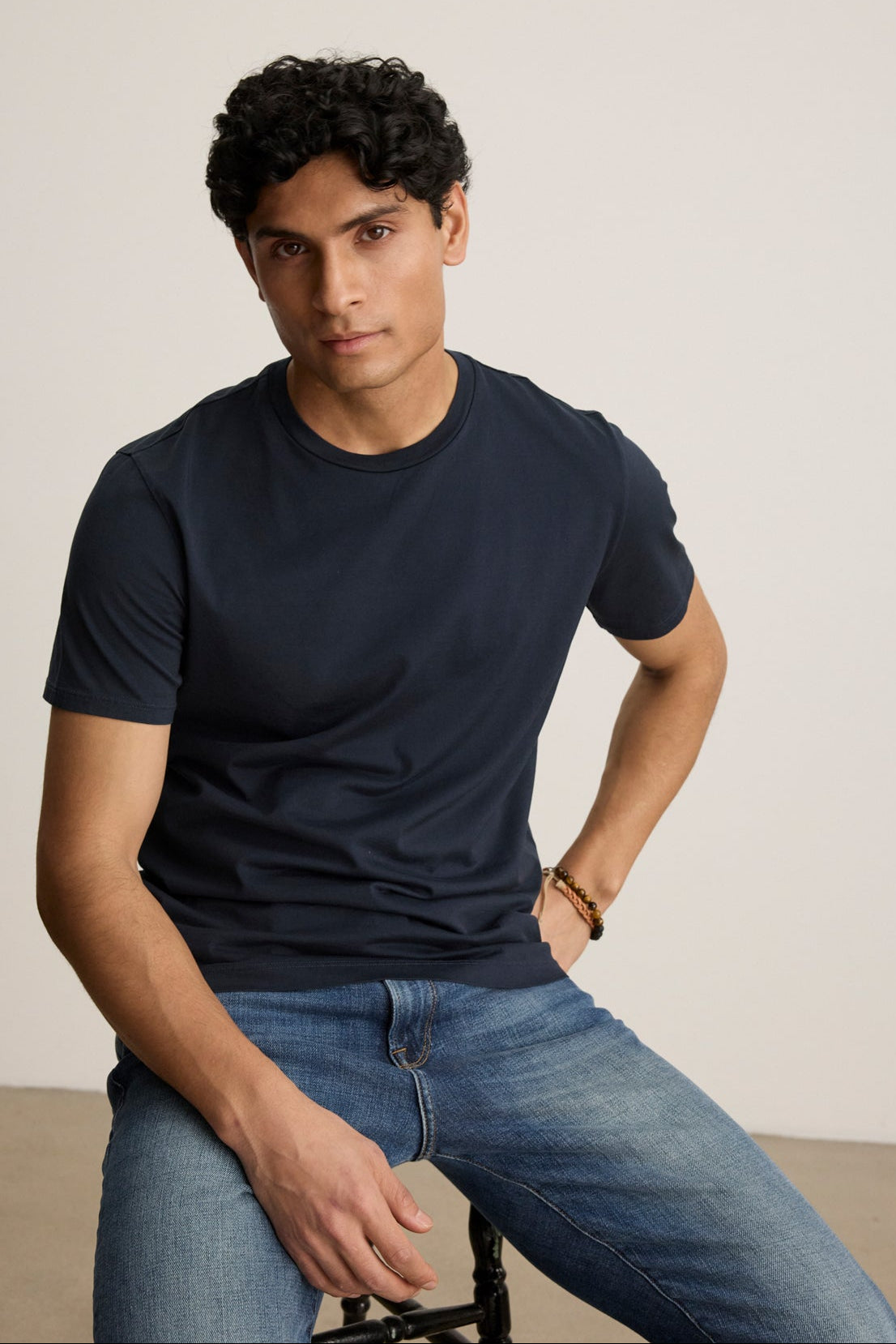 A man with short, curly black hair sits on a stool against a light background, wearing the Velvet by Graham & Spencer FRANKIE TEE in navy blue with a crew neckline and blue jeans.-39450522157249