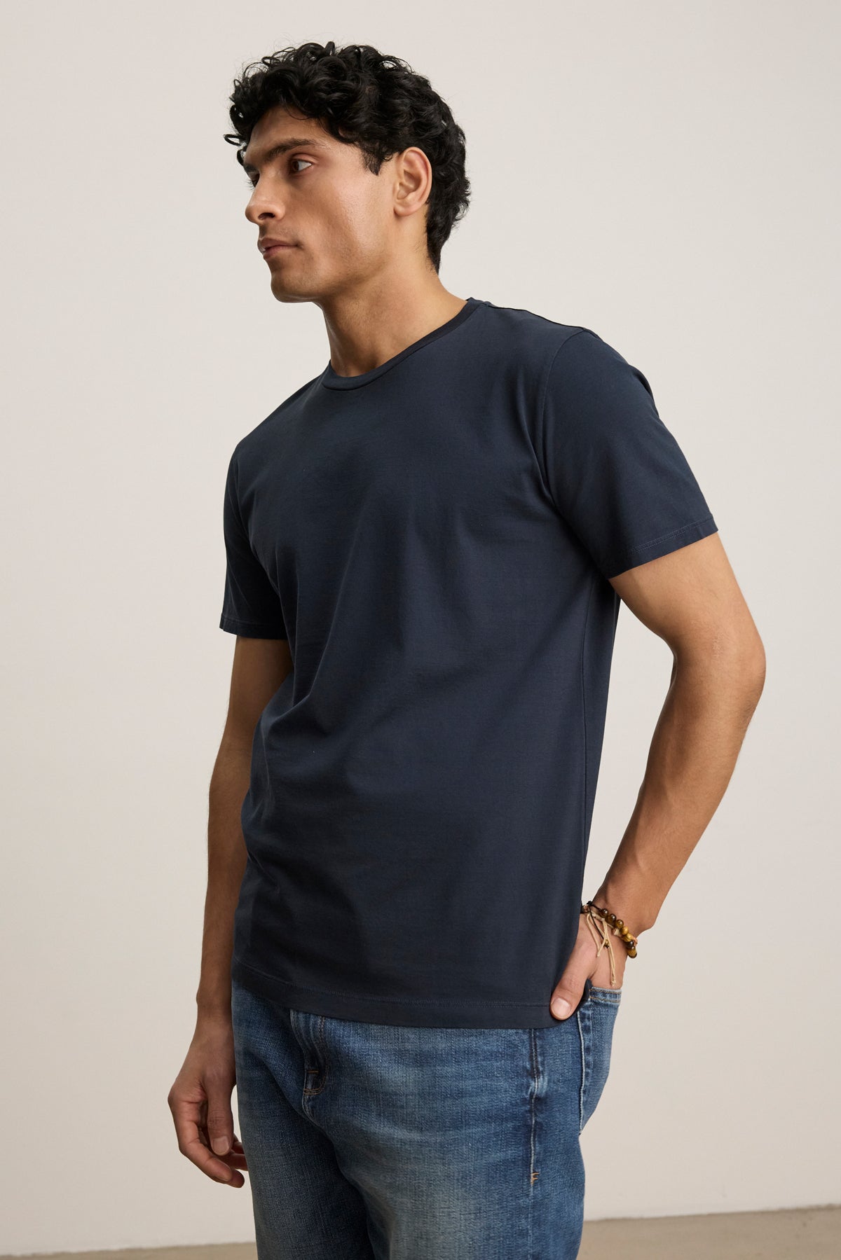 A man models the FRANKIE TEE by Velvet by Graham & Spencer—a navy blue, classic crewneck shirt—paired with blue jeans. He stands against a plain light background, looking to the side with one hand in his pocket.-39450522091713
