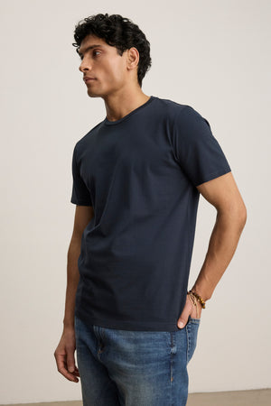 A man models the FRANKIE TEE by Velvet by Graham & Spencer—a navy blue, classic crewneck shirt—paired with blue jeans. He stands against a plain light background, looking to the side with one hand in his pocket.