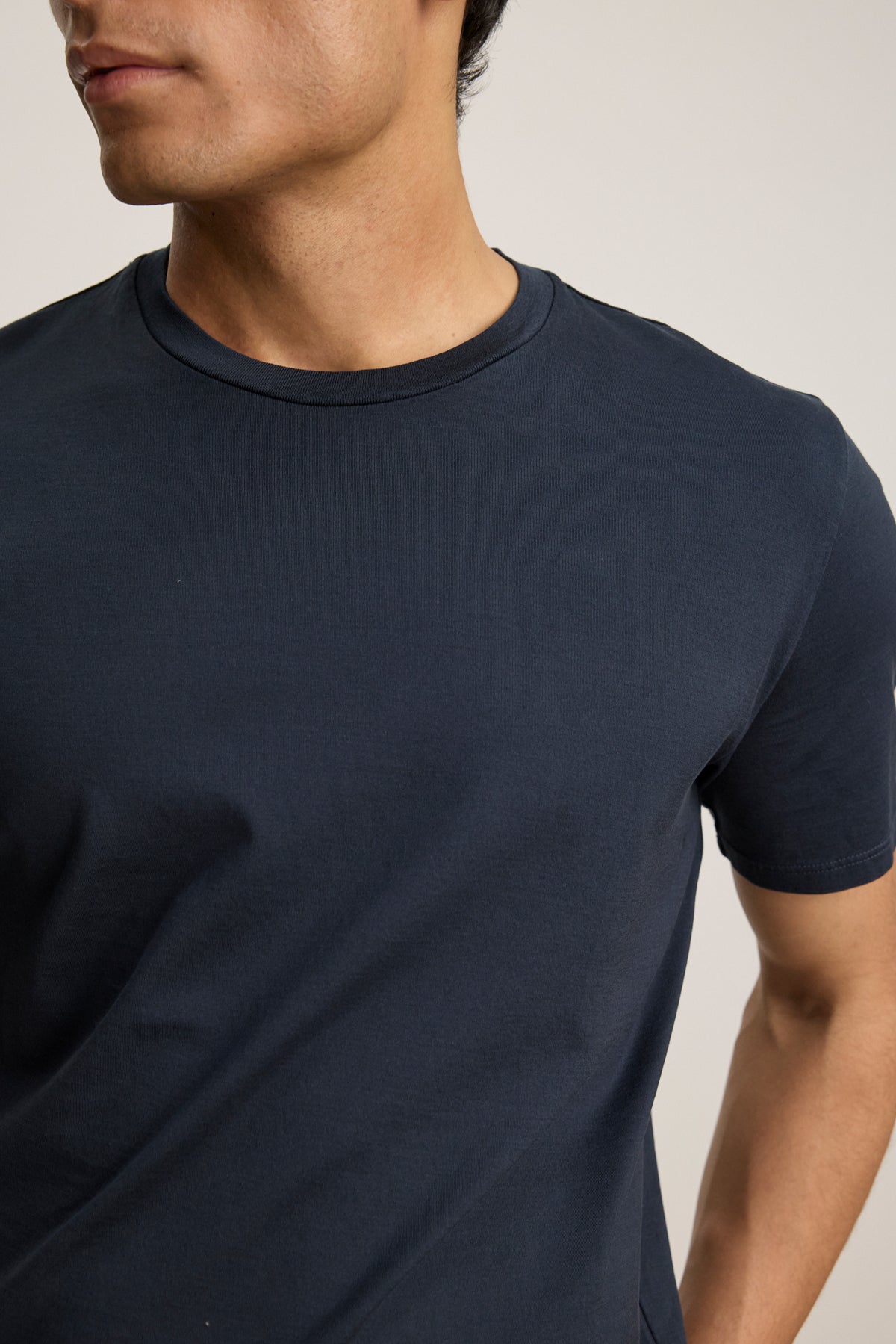 A person wears the FRANKIE TEE by Velvet by Graham & Spencer—a plain navy blue short-sleeve t-shirt with a classic crew neckline—shown from shoulders to mid-torso, facing slightly to the side.-39450522058945