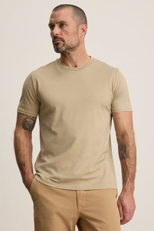 A man with short hair and a beard wears the FRANKIE TEE by Velvet by Graham & Spencer—a beige mercerized cotton t-shirt with a classic crew neckline—paired with tan pants, standing hands in pockets against a plain background.