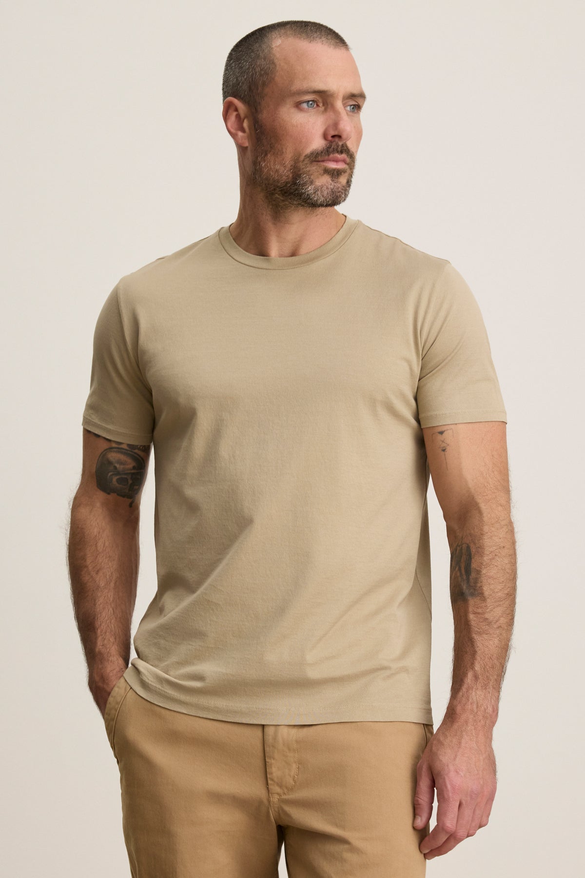 A man with short hair and a beard models the FRANKIE TEE by Velvet by Graham & Spencer—a beige crew neck t-shirt—paired with tan pants, standing hands-in-pockets against a plain backdrop; a versatile essential for any wardrobe.-39450521993409