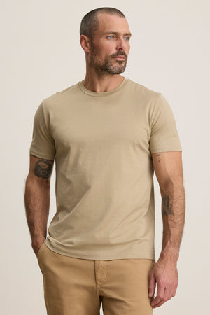 A man with short hair and a beard models the FRANKIE TEE by Velvet by Graham & Spencer—a beige crew neck t-shirt—paired with tan pants, standing hands-in-pockets against a plain backdrop; a versatile essential for any wardrobe.