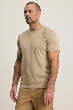 Man with short hair and beard wears the FRANKIE TEE by Velvet by Graham & Spencer—a beige mercerized cotton crew neck t-shirt—paired with tan pants, posing against a plain light background. A versatile wardrobe staple.