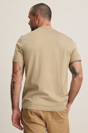 A man with short hair and tattoos stands facing away, wearing Velvet by Graham & Spencer's FRANKIE TEE in beige mercerized cotton with a classic crew neckline and light brown pants, set against a neutral background.