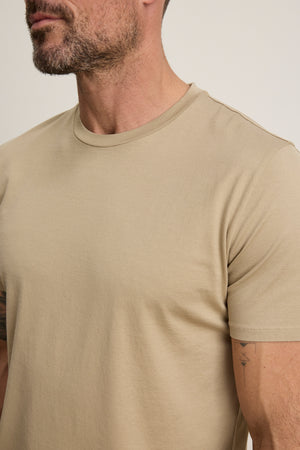 A man with a short beard models the FRANKIE TEE by Velvet by Graham & Spencer, a plain beige t-shirt with a classic crew neck, shown from shoulders to mid-torso against a neutral background.