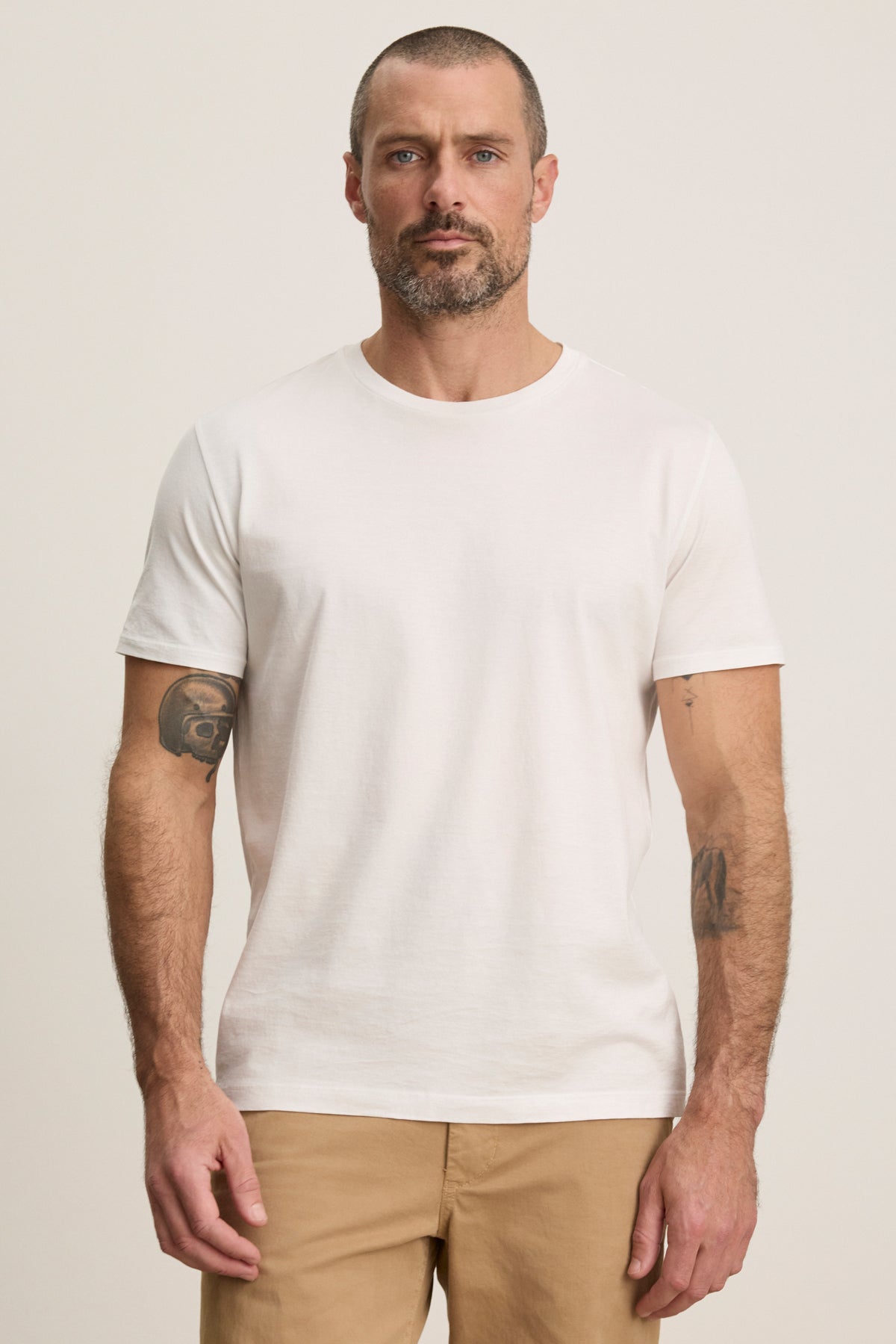 A man with short hair and a beard wears the Velvet by Graham & Spencer FRANKIE TEE, a classic white crewneck T-shirt, paired with beige pants, standing facing forward against a neutral background.-39450521862337