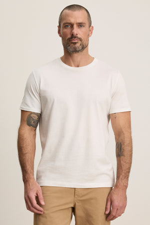 A man with short hair and a beard wears the Velvet by Graham & Spencer FRANKIE TEE, a classic white crewneck T-shirt, paired with beige pants, standing facing forward against a neutral background.