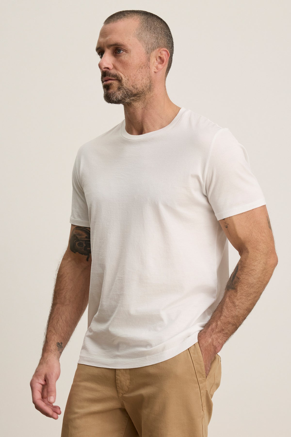 A man with short hair and a beard stands against a plain background, wearing the FRANKIE TEE by Velvet by Graham & Spencer—a white mercerized cotton crewneck paired with light brown pants for timeless, versatile style.-39450521796801