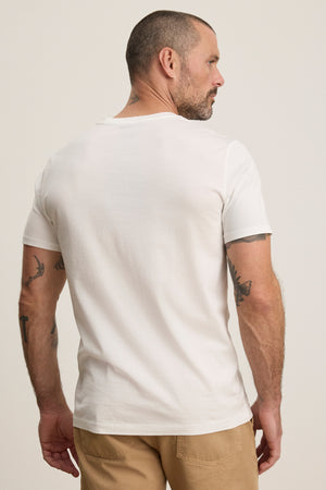 A man with short hair and tattoos stands with his back to the camera, wearing the Velvet by Graham & Spencer FRANKIE TEE in plain white and beige pants against a neutral background.