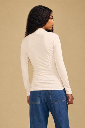 A woman with long curly hair stands with her back to the camera against a beige background, wearing Velvet by Graham & Spencer’s fitted, cream MERI TEE—a long-sleeve top with a flattering v-neckline—paired with blue jeans.