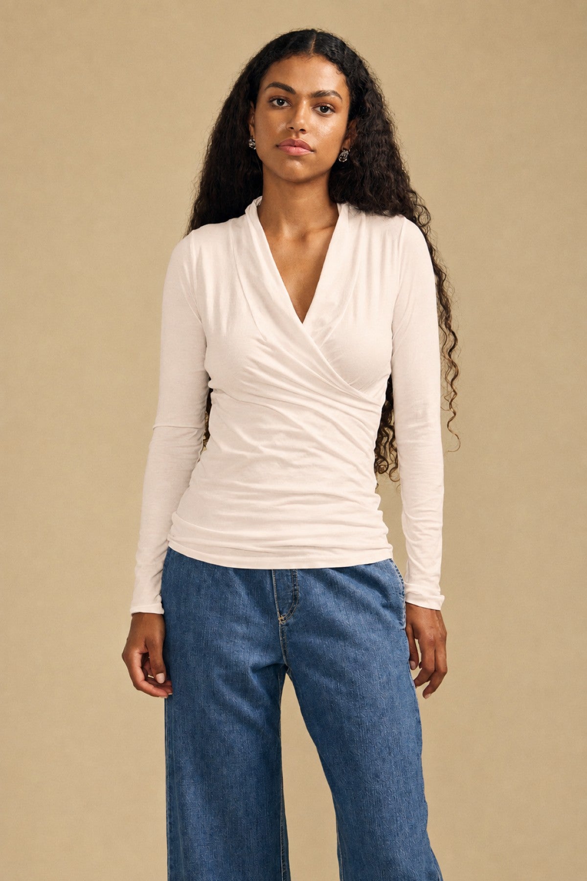 A woman with long curly hair wears the MERI TEE by Velvet by Graham & Spencer—a light wrap-style, long sleeve top with a flattering fitted silhouette—paired with blue wide-leg jeans, standing against a plain beige background.-41960122024129