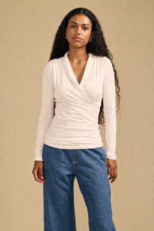 A woman with long curly hair wears the MERI TEE by Velvet by Graham & Spencer—a light wrap-style, long sleeve top with a flattering fitted silhouette—paired with blue wide-leg jeans, standing against a plain beige background.