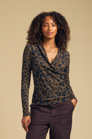 A woman stands against a beige background wearing the MERIANE TEE by Velvet by Graham & Spencer—a fitted silhouette, long-sleeve brown and black leopard print top—paired with dark brown pants, her hand in one pocket.