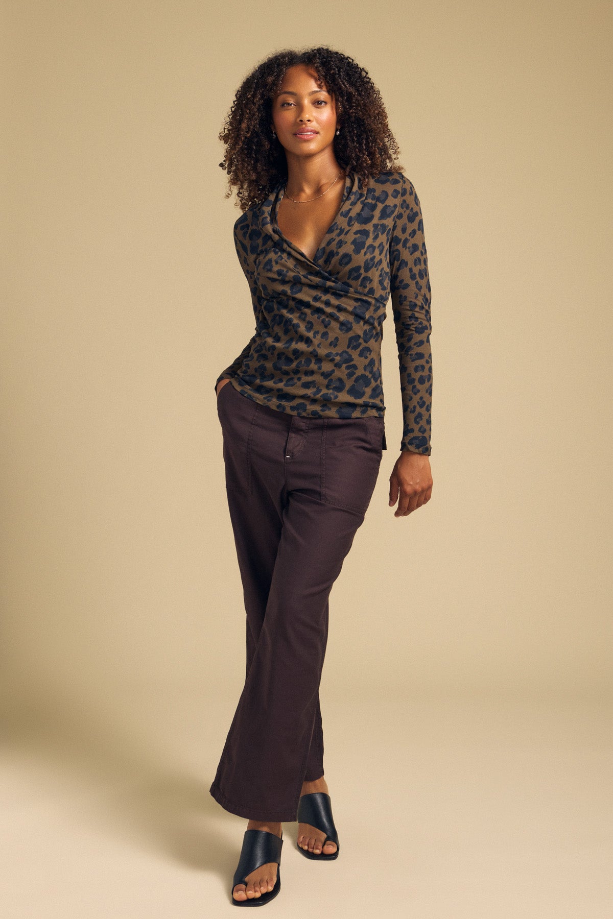 A woman stands against a beige background wearing the MERIANE TEE by Velvet by Graham & Spencer—a leopard print top with a fitted silhouette—paired with dark brown pants and black open-toe shoes.