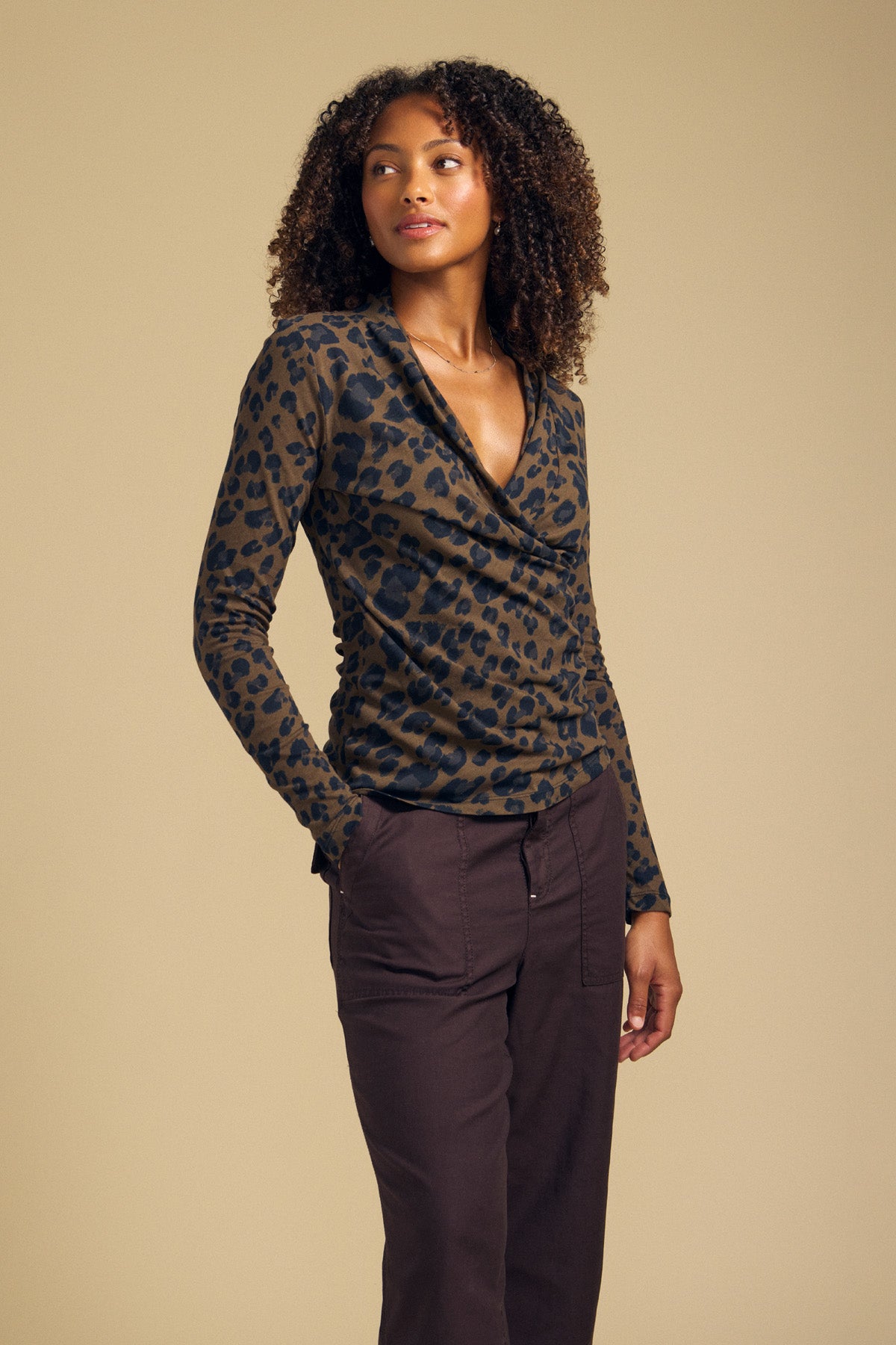 A woman stands against a plain background wearing the Velvet by Graham & Spencer MERIANE TEE—a fitted, long-sleeve leopard print cowl neck top crafted from soft cotton jersey—paired with high-waisted, dark brown pants, gazing off to the side.