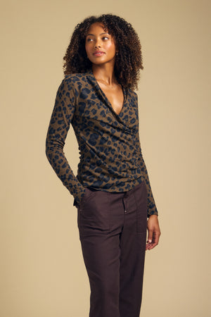 A woman stands against a plain background wearing the Velvet by Graham & Spencer MERIANE TEE—a fitted, long-sleeve leopard print cowl neck top crafted from soft cotton jersey—paired with high-waisted, dark brown pants, gazing off to the side.