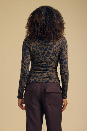 A person stands with their back to the camera, wearing the Velvet by Graham & Spencer MERIANE TEE—featuring a fitted silhouette and leopard print—paired with dark pants, set against a plain beige background.