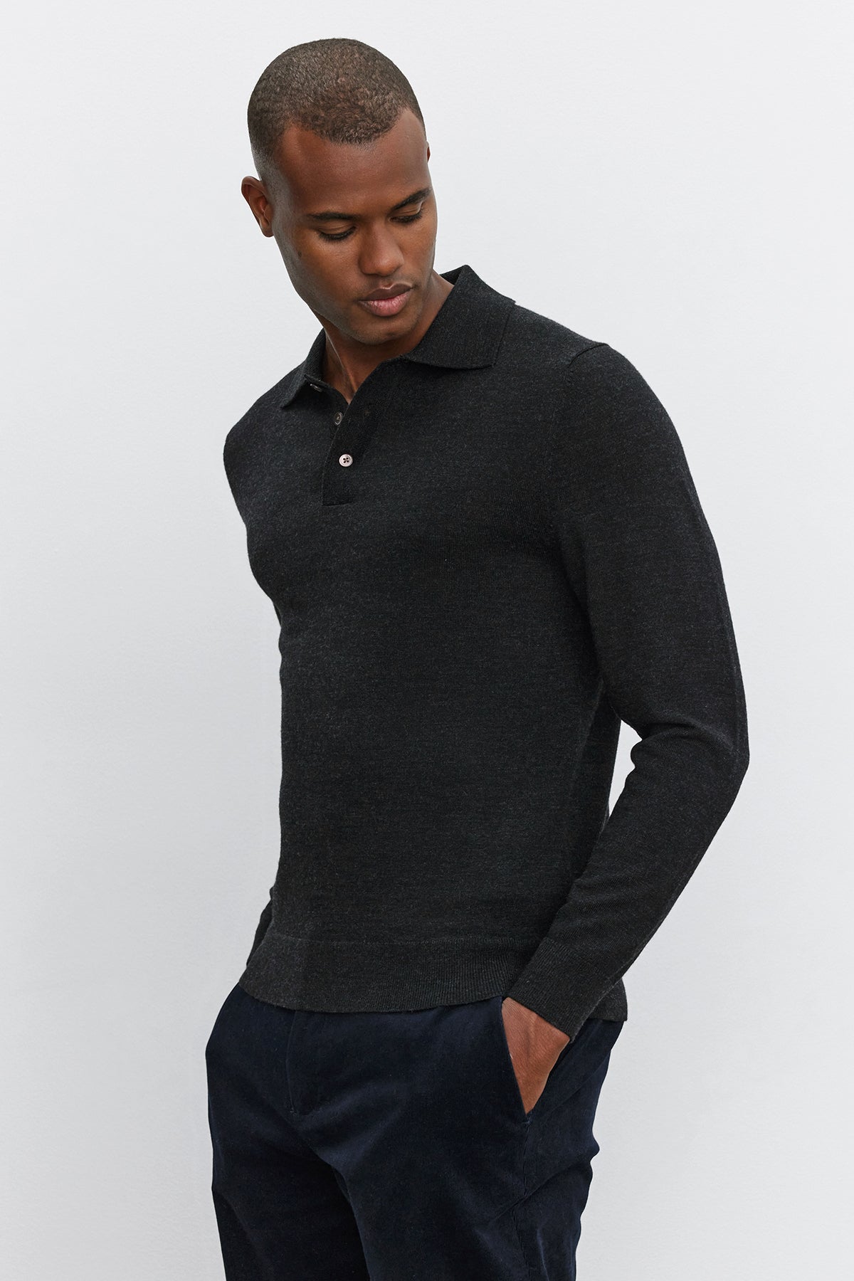   A person with short hair wearing a long-sleeved, dark ARTHUR SWEATER POLO by Velvet by Graham & Spencer and dark pants, looking down with hands in pockets. 