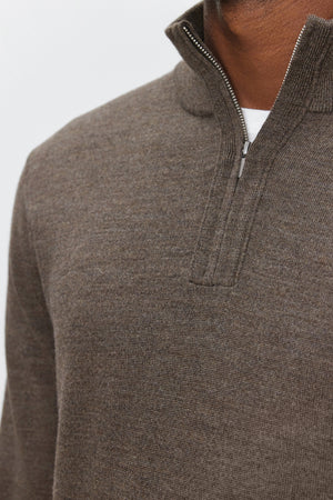 Close-up of a person wearing the FLETCHER QUARTER-ZIP SWEATER by Velvet by Graham & Spencer in brown merino wool, revealing a white shirt underneath. The background is plain and light-colored.