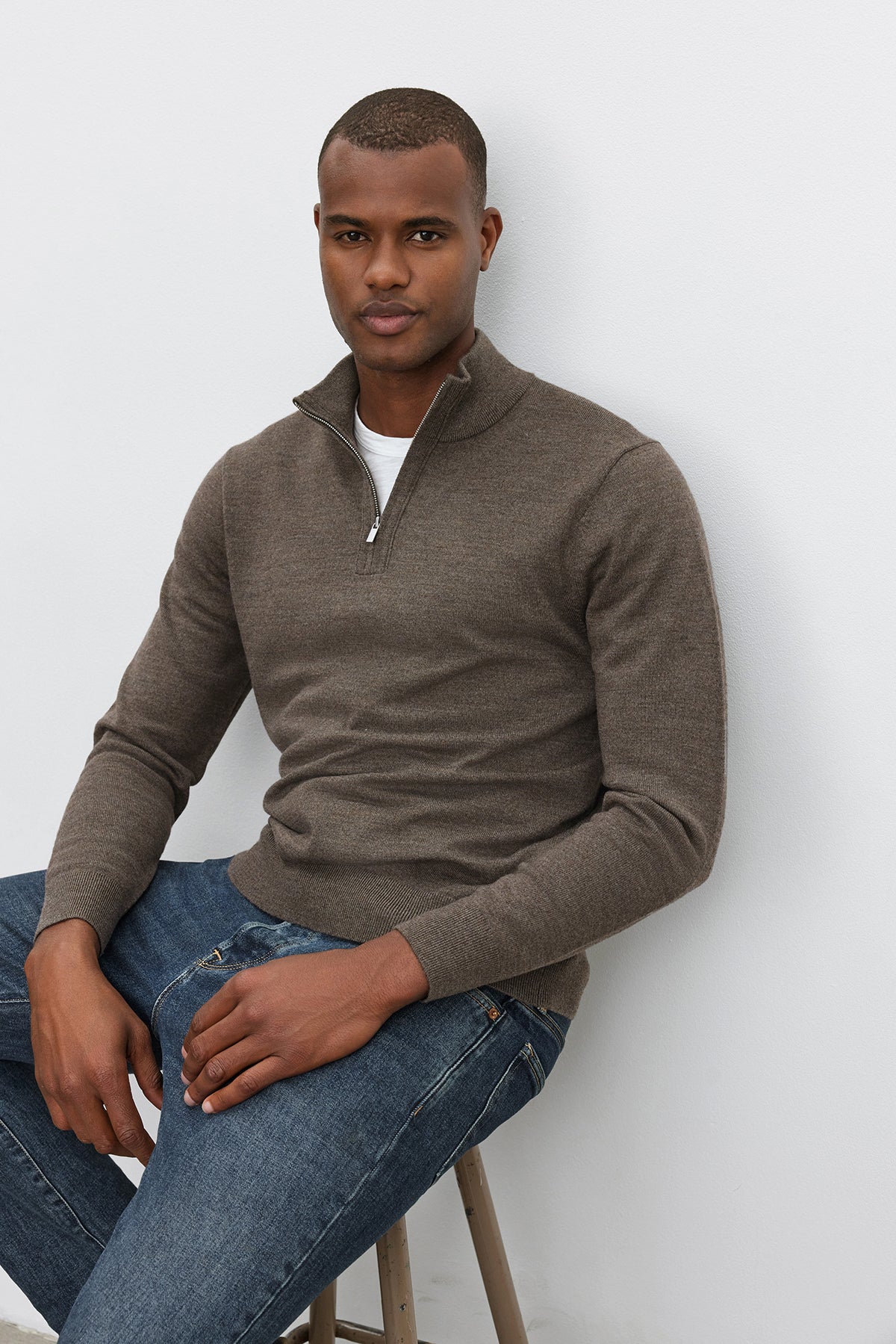   A person wearing the FLETCHER QUARTER-ZIP SWEATER from Velvet by Graham & Spencer in brown over a white shirt and blue jeans sits on a stool against a plain white background. 