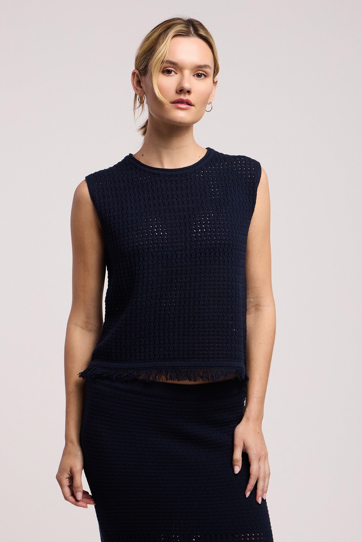 A woman faces the camera against a plain light background, wearing the Velvet by Graham & Spencer COVA SWEATER—a sleeveless, textured navy mesh-stitch top—paired with a matching skirt.