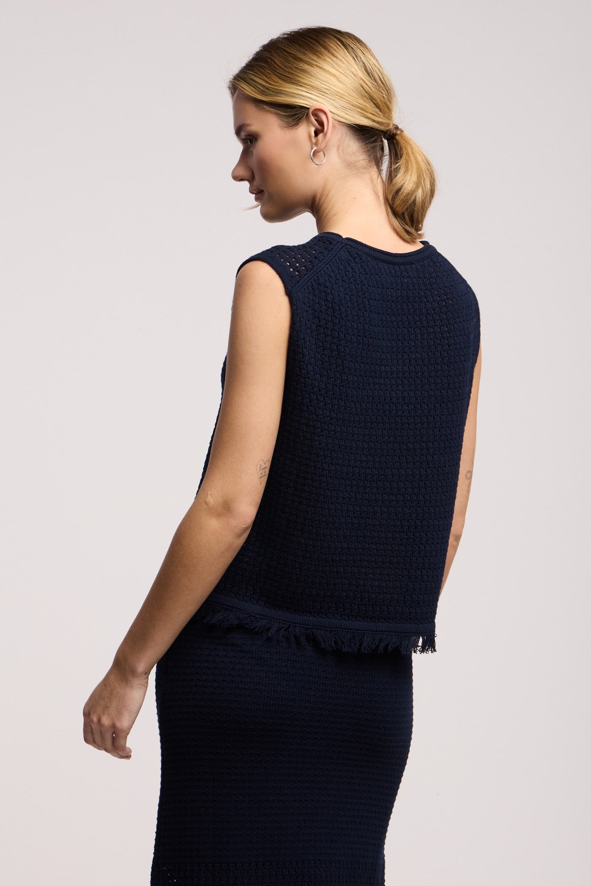 A woman with blonde hair in a low ponytail models the COVA SWEATER by Velvet by Graham & Spencer, styled with a matching skirt and photographed from the side and back against a plain background.
