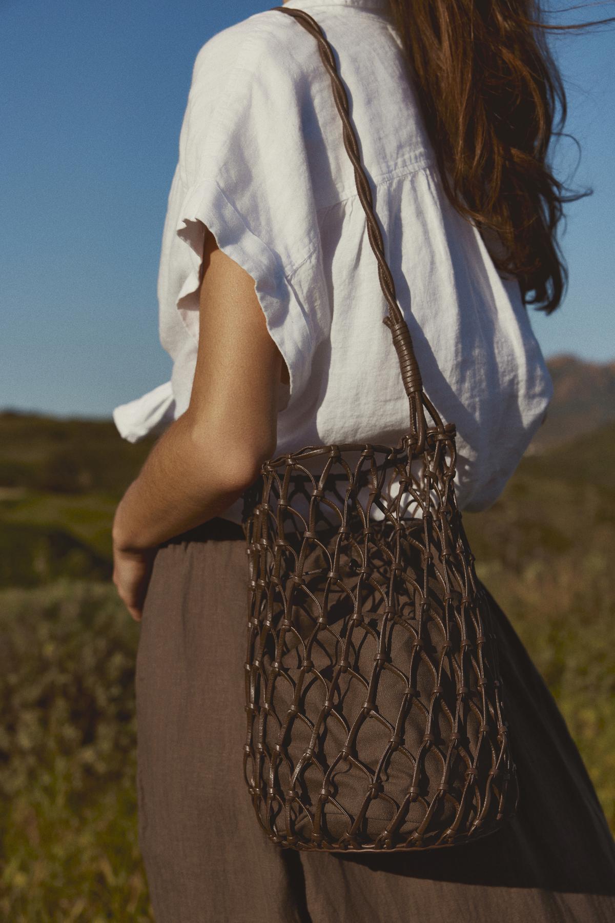   Woman in a white blouse and brown skirt holding a Velvet by Graham & Spencer mesh tote bag with a mesh design, standing in a field under clear skies. 