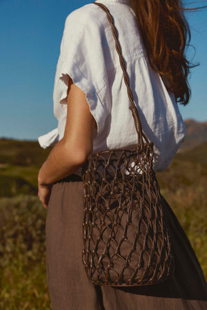 Woman in a white blouse and brown skirt holding a Velvet by Graham & Spencer mesh tote bag with a mesh design, standing in a field under clear skies.