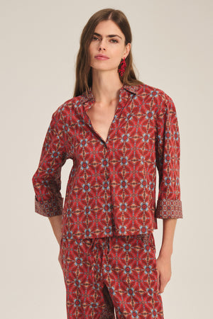 A woman wears the Velvet by Graham & Spencer MICAH SHIRT, a red patterned button-up pajama top with matching long pants, standing against a plain background.