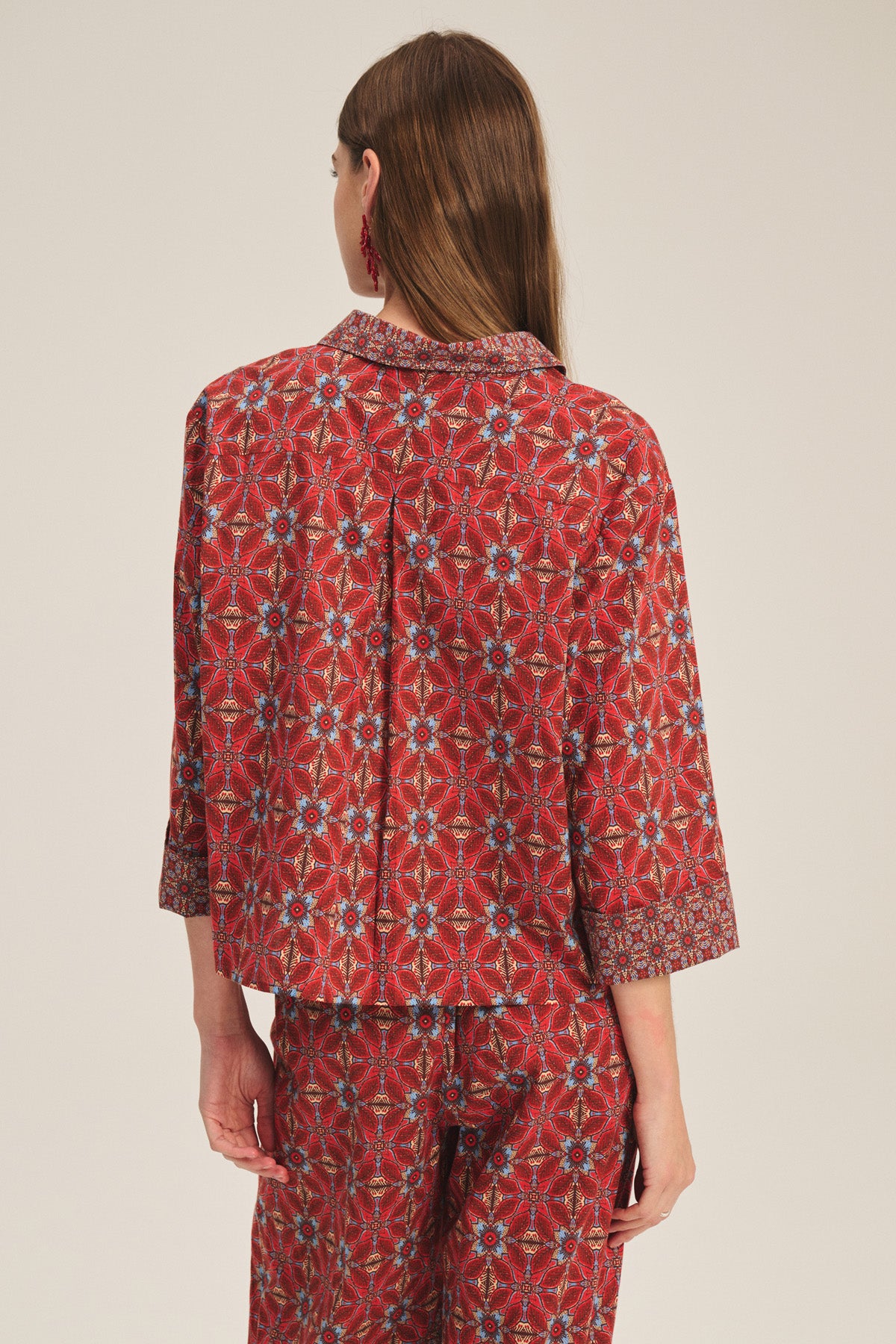 A woman stands facing away, wearing the Velvet by Graham & Spencer MICAH SHIRT—a red patterned top with blue and white geometric designs.-41436010643649