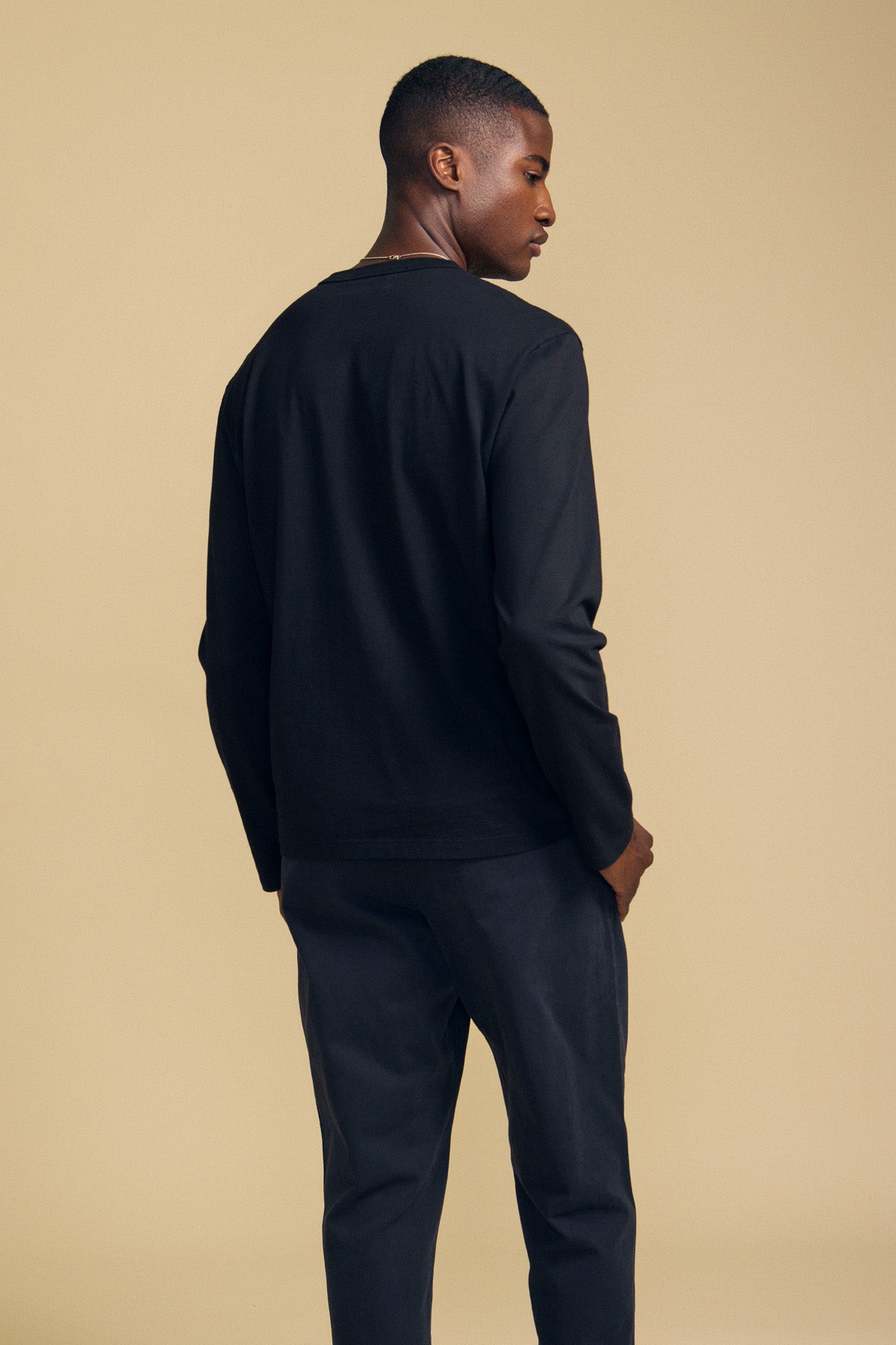 A person stands facing away, wearing the black ANTONIO TEE by Velvet by Graham & Spencer and black pants against a beige background.