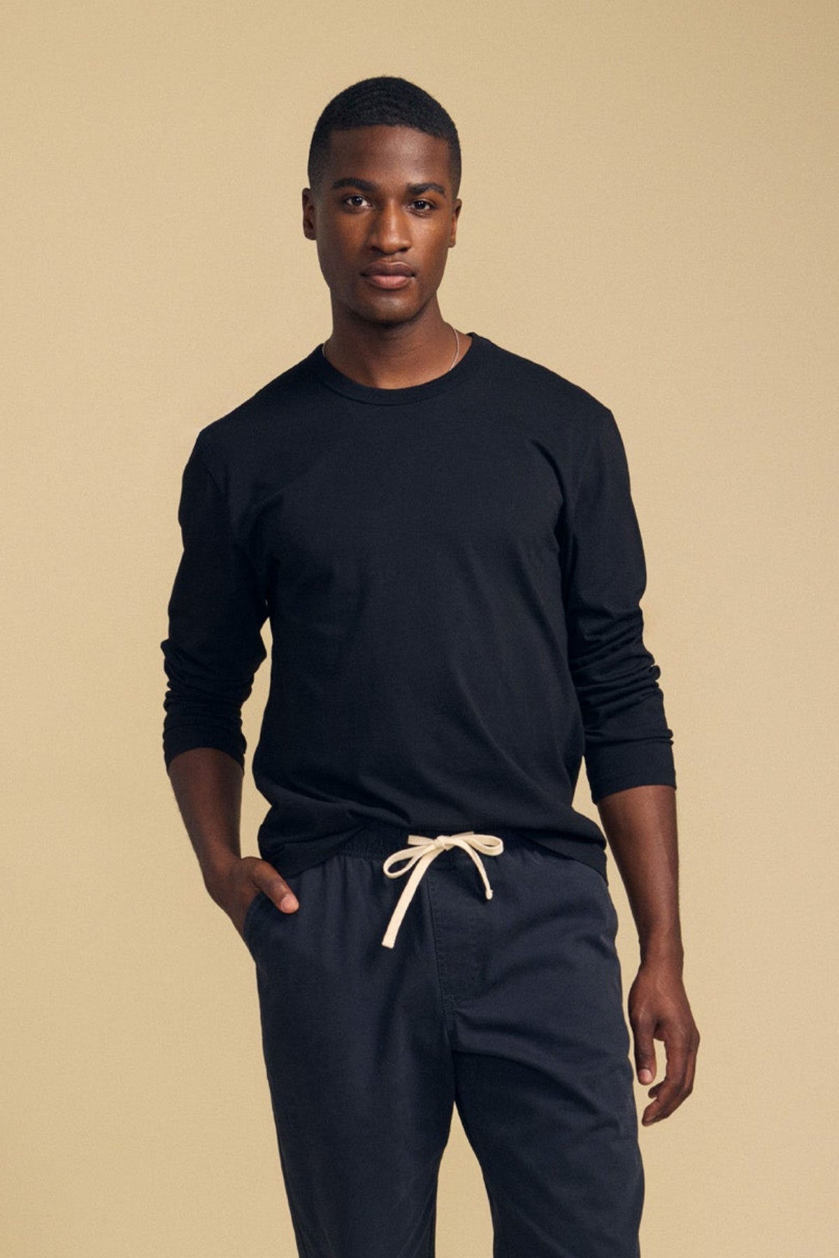 A person stands against a beige background wearing the Velvet by Graham & Spencer ANTONIO TEE, a black cotton jersey long sleeve, paired with dark pants featuring a white drawstring, with one hand in their pocket.