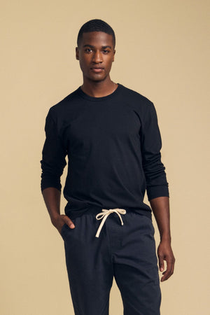 A person stands against a beige background wearing the Velvet by Graham & Spencer ANTONIO TEE, a black cotton jersey long sleeve, paired with dark pants featuring a white drawstring, with one hand in their pocket.