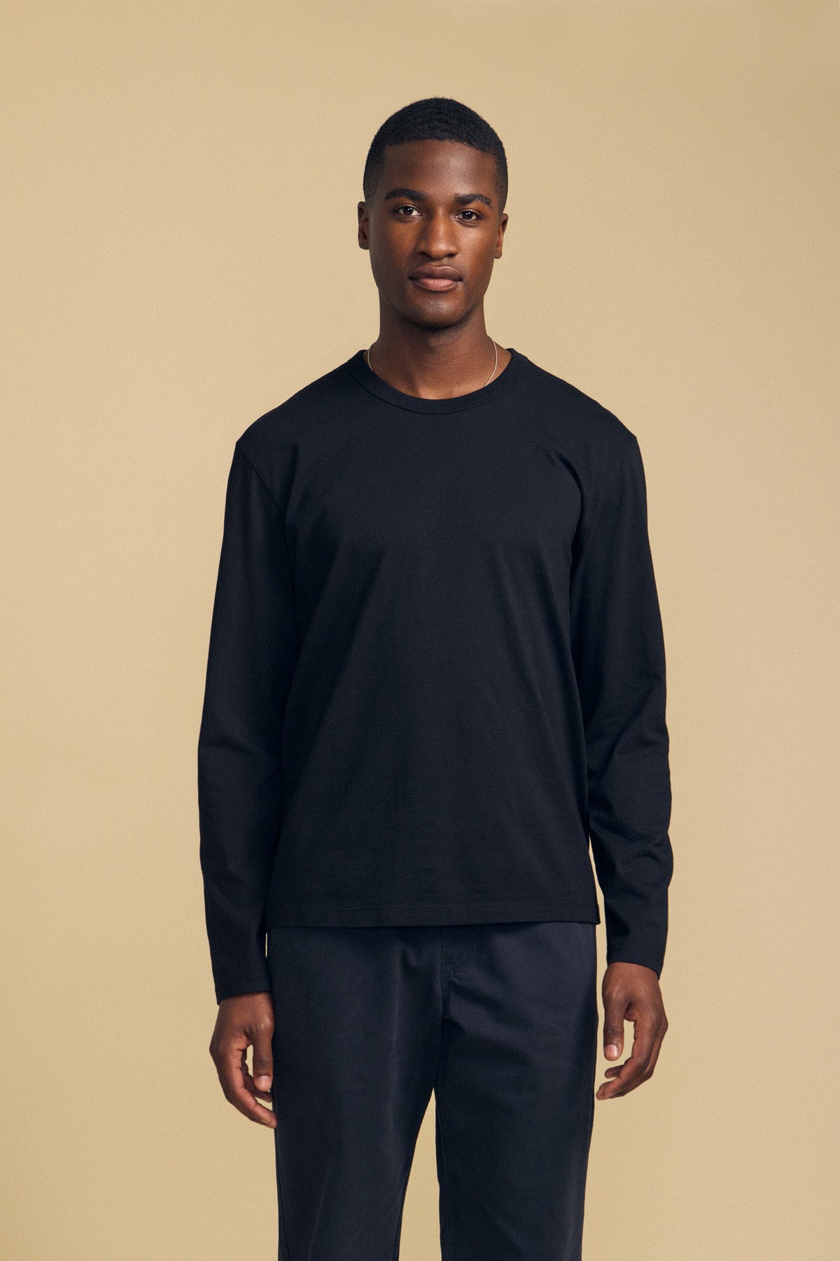 A man stands against a beige background, wearing the ANTONIO TEE from Velvet by Graham & Spencer—a relaxed fit black long sleeve crafted from soft cotton jersey—paired with matching black pants, facing the camera with a neutral expression.-42092436848833