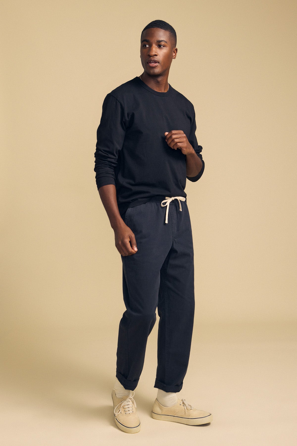 A man stands against a beige background wearing the Velvet by Graham & Spencer ANTONIO TEE, a black cotton jersey long sleeve, paired with dark drawstring pants and beige sneakers. He gazes to the side with one hand partially raised, showcasing a relaxed fit style.-42092437569729