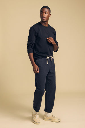 A man stands against a beige background wearing the Velvet by Graham & Spencer ANTONIO TEE, a black cotton jersey long sleeve, paired with dark drawstring pants and beige sneakers. He gazes to the side with one hand partially raised, showcasing a relaxed fit style.