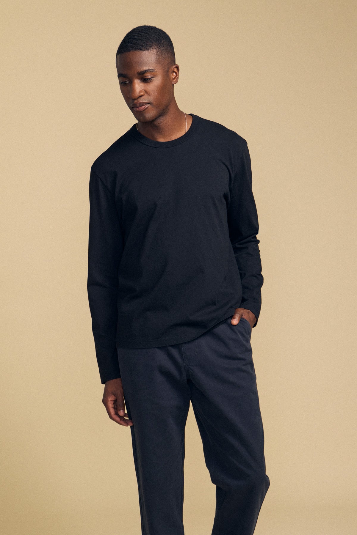 A person stands against a beige background, wearing the ANTONIO TEE by Velvet by Graham & Spencer—a relaxed fit long sleeve tee crafted from black cotton jersey—paired with black pants, looking downward with one hand in a pocket.