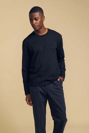 A person stands against a beige background, wearing the ANTONIO TEE by Velvet by Graham & Spencer—a relaxed fit long sleeve tee crafted from black cotton jersey—paired with black pants, looking downward with one hand in a pocket.