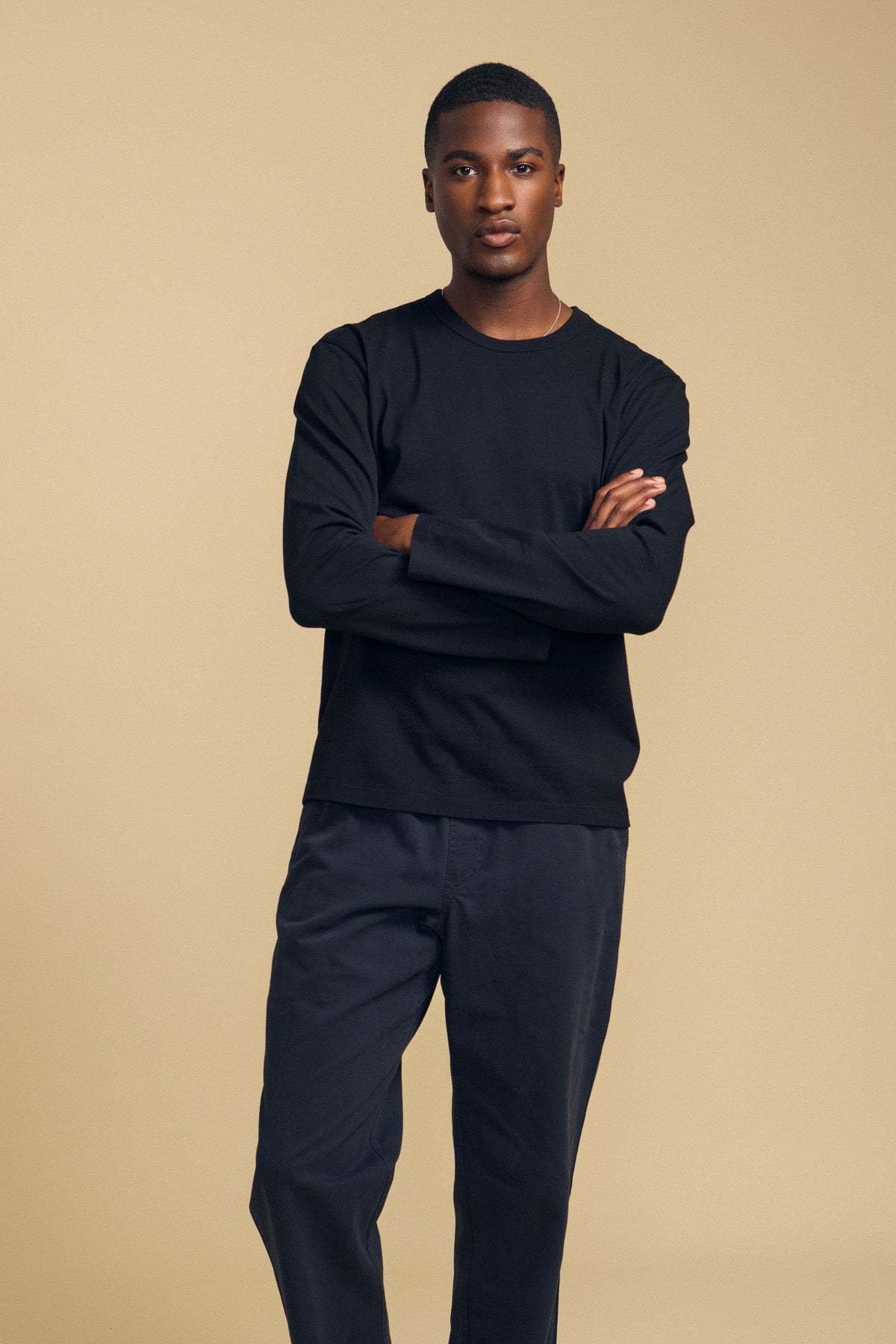 A man stands with arms crossed, wearing the Velvet by Graham & Spencer ANTONIO TEE in black—a relaxed fit, long-sleeve cotton jersey shirt—paired with matching black pants against a plain beige background. The cotton jersey fabric lends the look a comfortable and effortless appeal.