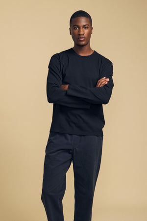 A man stands with arms crossed, wearing the Velvet by Graham & Spencer ANTONIO TEE in black—a relaxed fit, long-sleeve cotton jersey shirt—paired with matching black pants against a plain beige background. The cotton jersey fabric lends the look a comfortable and effortless appeal.