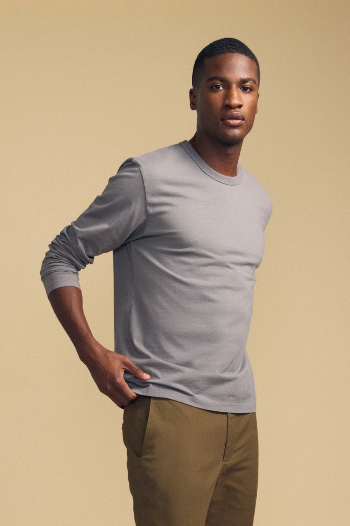 A man wearing the ANTONIO TEE by Velvet by Graham & Spencer—a light gray cotton jersey long sleeve—and tan pants stands against a plain beige background, looking forward with a neutral expression.