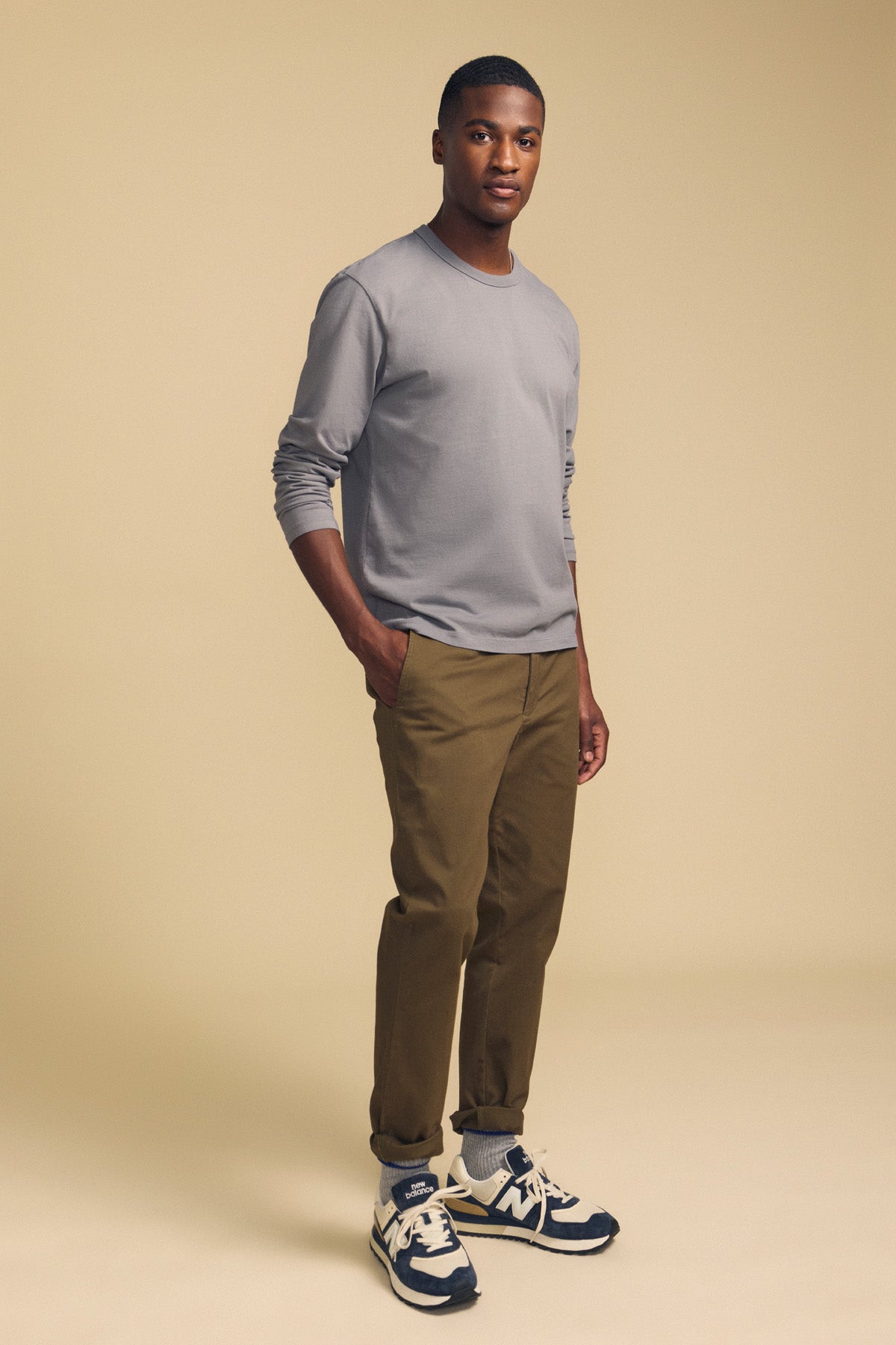 A man stands against a beige background with his hands in his pockets and a neutral expression, wearing the Velvet by Graham & Spencer ANTONIO TEE—a relaxed fit grey cotton jersey long sleeve—paired with khaki pants and black and white sneakers.-42092438094017